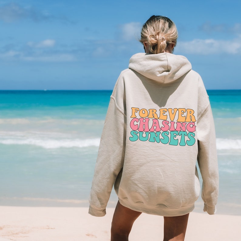 May include: A beige hoodie with the text "FOREVER CHASING SUNSETS" in colorful, retro-style lettering on the back. The hoodie is worn by a person standing on a beach.