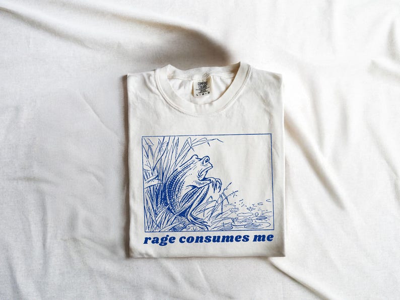 May include: White t-shirt with a blue illustration of a frog in a pond. The text "rage consumes me" is printed below the image.