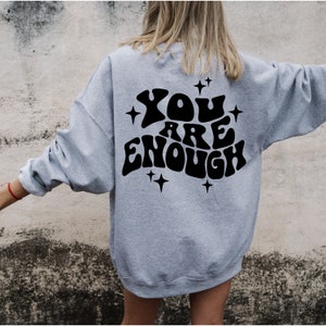 Puede incluir: Una sudadera gris con el texto "YOU ARE ENOUGH" en negro, con letras de estilo retro y estrellas alrededor.