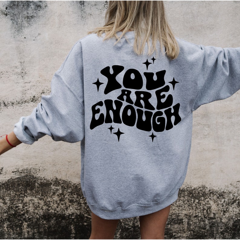 Puede incluir: Una sudadera gris con el texto "YOU ARE ENOUGH" en negro, con letras de estilo retro y estrellas alrededor.