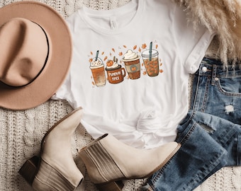 Puede incluir: Camiseta blanca con un gráfico de cuatro bebidas de café con temática otoñal. Las bebidas están decoradas con hojas de otoño, calabazas y crema batida. El texto "Pumpkin Spice" está escrito en la camiseta.