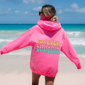 May include: Pink hoodie with the text "FOREVER CHASING SUNSETS" in a retro font on the back.