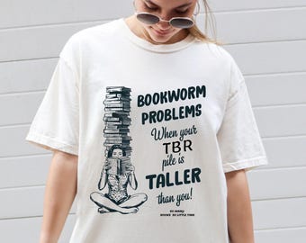 May include: White t-shirt with a black and white illustration of a woman reading a book. The text on the shirt reads "Bookworm Problems When your TBR pile is Taller than you! So many books so little time."
