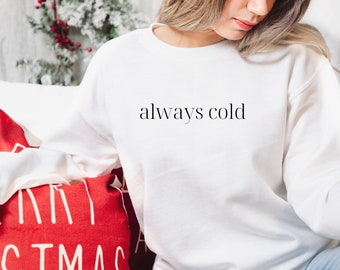 Puede incluir: Una sudadera blanca con el texto "always cold" impreso en la parte delantera. La sudadera la lleva puesta una persona sentada en un sofá.