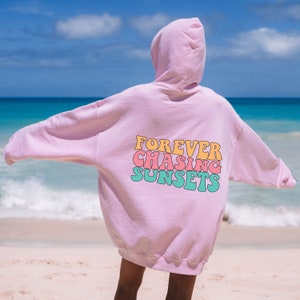 May include: Pink oversized hoodie with the text "FOREVER CHASING SUNSETS" in colorful retro font on the back. The hoodie is worn by a person standing on a beach.