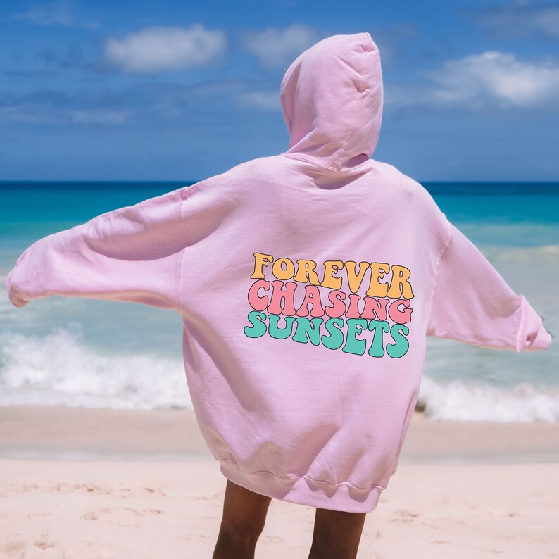 May include: Pink oversized hoodie with the text "FOREVER CHASING SUNSETS" in colorful retro font on the back. The hoodie is worn by a person standing on a beach.