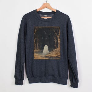 Puede incluir: Una sudadera gris oscuro jaspeada con un estampado gr&aacute;fico de un fantasma blanco en un entorno de bosque. El fantasma est&aacute; de pie en un charco de agua con lluvia cayendo a su alrededor.