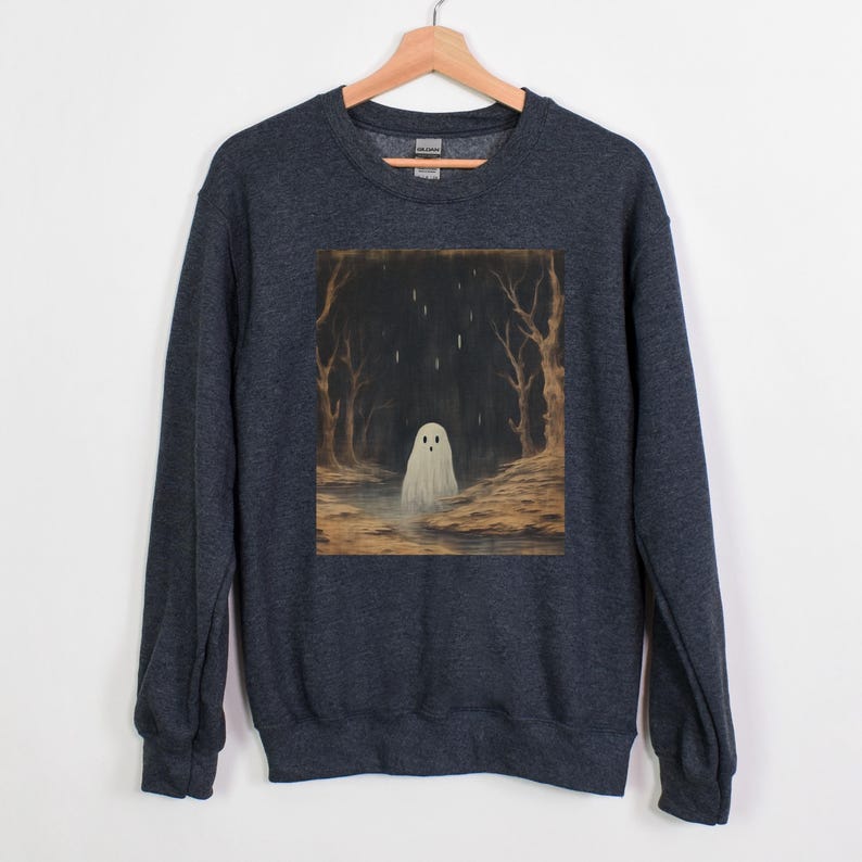 Puede incluir: Una sudadera gris oscuro jaspeada con un estampado gr&aacute;fico de un fantasma blanco en un entorno de bosque. El fantasma est&aacute; de pie en un charco de agua con lluvia cayendo a su alrededor.