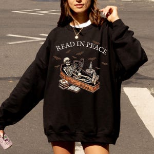 May include: Black sweatshirt with a skeleton reading a book in a coffin. The text "Read in Peace" is printed on the front of the sweatshirt.