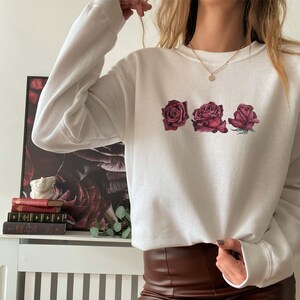 K&ouml;nnte beinhalten: Wei&szlig;er Pullover mit Rundhalsausschnitt und langen &Auml;rmeln, auf dem vorne drei burgunderrote Rosen aufgedruckt sind.