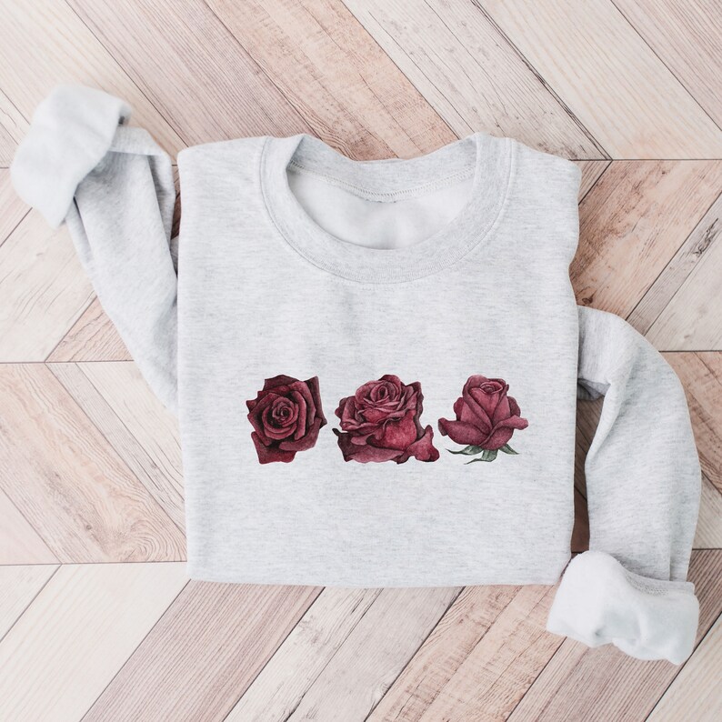 K&ouml;nnte beinhalten: Ein grauer Pullover mit einem Design aus drei burgunderfarbenen Rosen auf der Vorderseite.