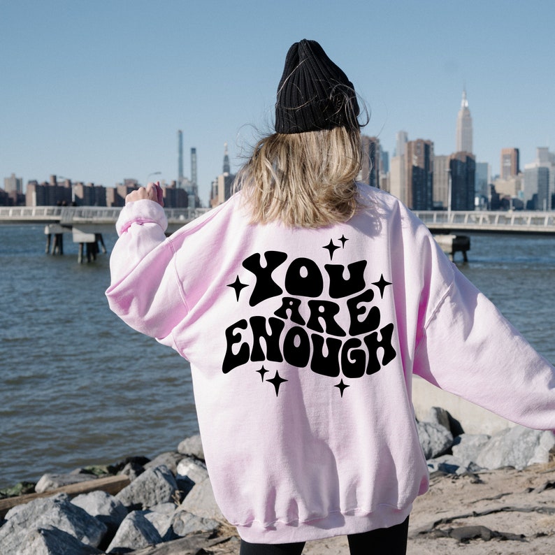 Puede incluir: Una sudadera rosa con el texto "YOU ARE ENOUGH" en negro, con letras de estilo retro. La sudadera la lleva puesta una persona de pie frente a un horizonte urbano.