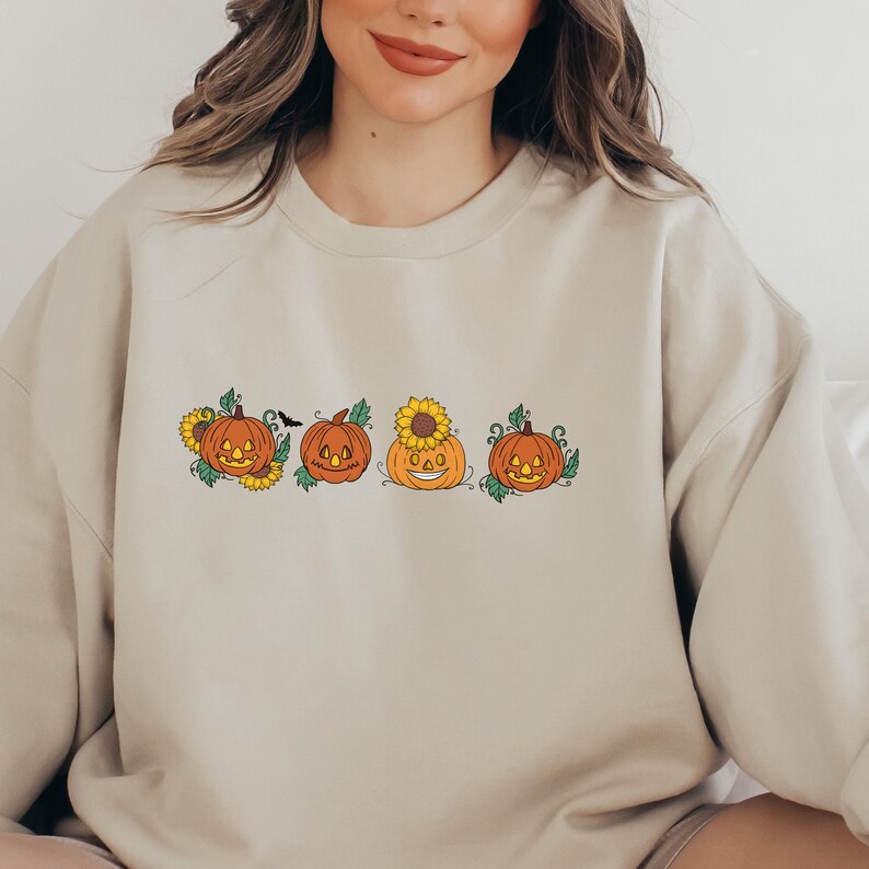 May include: A beige sweatshirt featuring a design of four smiling jack-o'-lantern pumpkins with green vines and sunflowers.