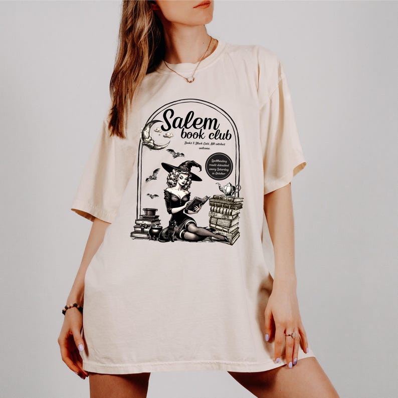 Puede incluir: Una camiseta de color crema con una ilustraci&oacute;n en blanco y negro de una bruja leyendo un libro frente a una pila de libros. El texto "Salem book club" est&aacute; impreso en la camiseta.