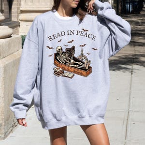 May include: Gray sweatshirt with a skeleton reading a book in a coffin. The text "Read in Peace" is printed on the front of the sweatshirt.