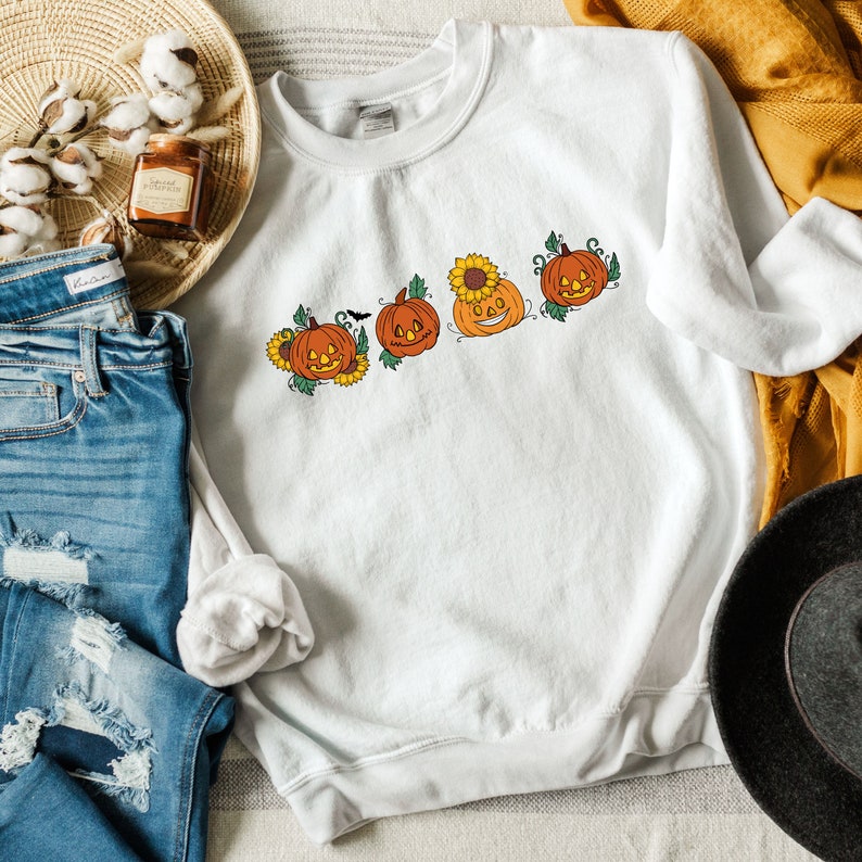 May include: White sweatshirt featuring a graphic of four orange pumpkins with jack-o'-lantern faces and green stems. The pumpkins are arranged in a row and have sunflowers behind them.