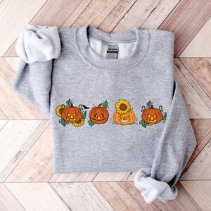 May include: A grey sweatshirt featuring a design of four orange pumpkins with smiling faces. The pumpkins are surrounded by green leaves and yellow sunflowers.