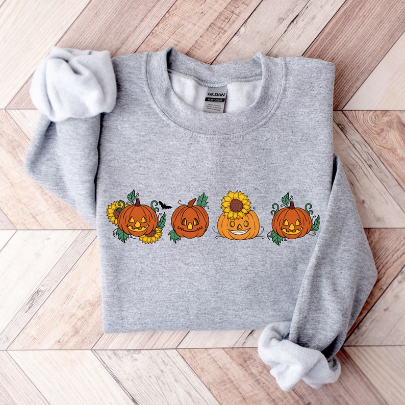 May include: A grey sweatshirt featuring a design of four orange pumpkins with smiling faces. The pumpkins are surrounded by green leaves and yellow sunflowers.