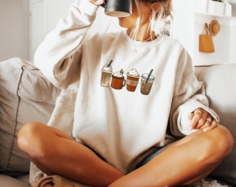 May include: A white sweatshirt with a graphic of four different coffee drinks. The drinks are all topped with whipped cream and have different flavors, including pumpkin spice and caramel.