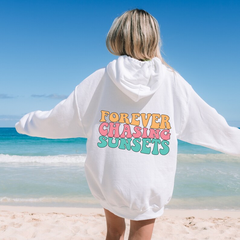 May include: White hoodie with a graphic print on the back that says "FOREVER CHASING SUNSETS" in a retro font with pink, orange, and blue colors. The hoodie is worn by a person standing on a beach.
