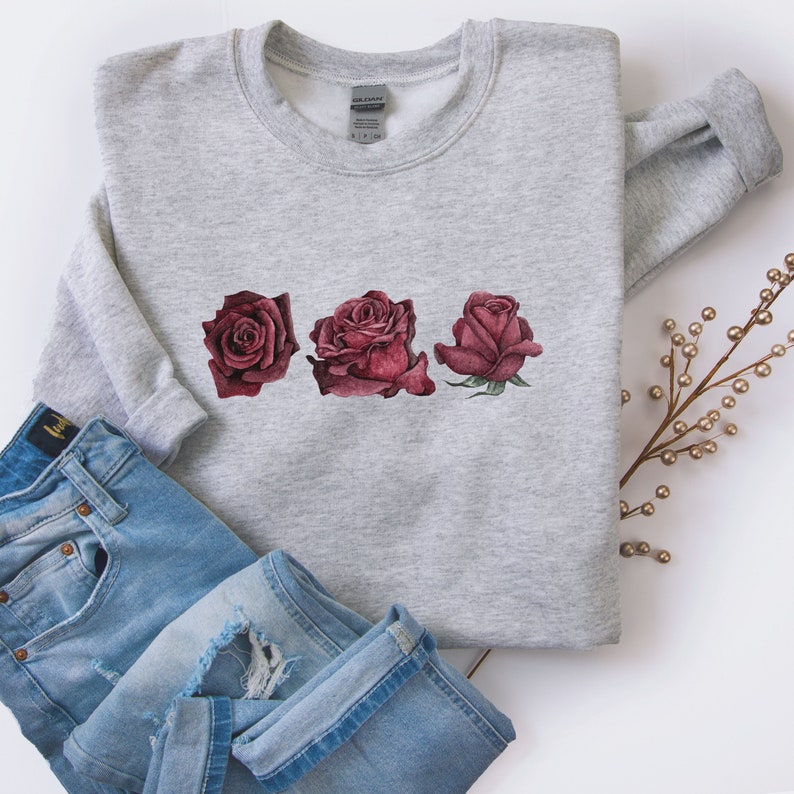 K&ouml;nnte beinhalten: Ein grauer Pullover mit einem Design aus drei roten Rosen auf der Vorderseite. Die Rosen sind in einer Reihe angeordnet und &uuml;berlappen sich leicht. Der Pullover ist gefaltet und auf einer wei&szlig;en Oberfl&auml;che abgelegt.