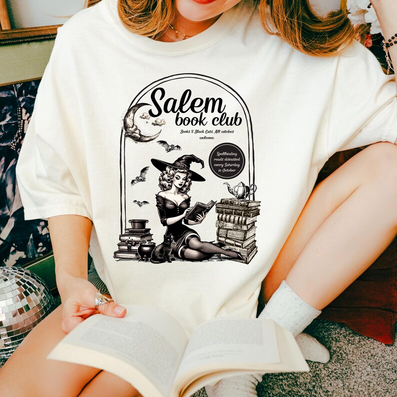 Puede incluir: Camiseta blanca con una ilustraci&oacute;n en blanco y negro de una bruja leyendo un libro frente a una pila de libros. El texto "Salem Book Club" est&aacute; por encima de la bruja. El texto "Books & Black Cats, All witches welcome" est&aacute; debajo de la bruja. El texto "Spellbinding monthly discussions every Saturday in October" est&aacute; a la derecha de la bruja.
