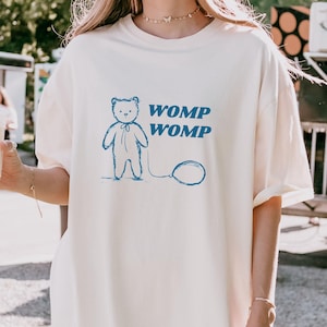 Może przedstawiać: Biały t-shirt z niebieską ilustracją pluszowego misia trzymającego balon. Tekst "WOMP WOMP" jest nadrukowany na niebiesko pod misiem.