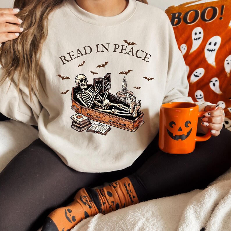 May include: A beige sweatshirt with the text "Read in Peace" and a skeleton reading a book in a coffin. The skeleton is wearing a black shirt and has a black skull. The coffin is brown and has a white candle on top. There are bats flying around the skeleton and coffin.