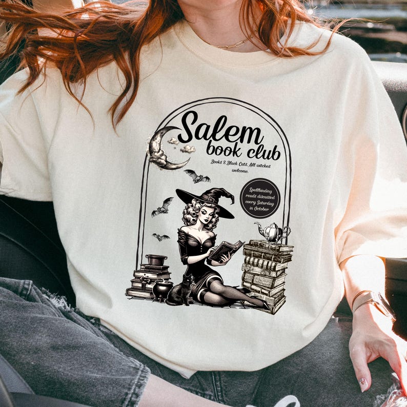 Puede incluir: Una camiseta blanca con un dise&ntilde;o gr&aacute;fico de una bruja leyendo un libro frente a una pila de libros. El gr&aacute;fico dice "Salem Book Club" y "Books & Black Cats, All witches welcome." El gr&aacute;fico tambi&eacute;n dice "Spellbinding reads distributed every Saturday in October."