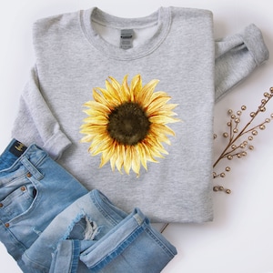 Könnte beinhalten: Ein graues Sweatshirt mit einem gelben Sonnenblumen-Grafikdruck. Die Sonnenblume hat eine braune Mitte und gelbe Blütenblätter. Das Sweatshirt ist gefaltet und auf einer weißen Oberfläche mit einer blauen Jeans gelegt.