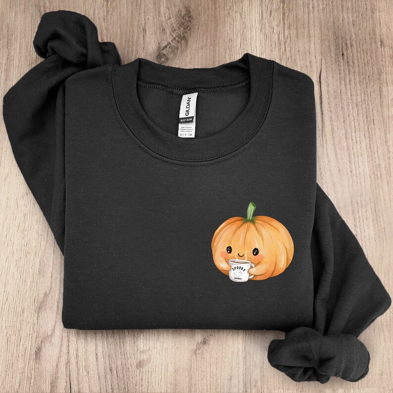 以下が含まれることがあります： コーヒーカップを持ったかわいいカボチャの漫画が描かれた黒いスウェットシャツ。カボチャは笑顔で、カップには「Spooky」と書かれた白いカップを持っています。