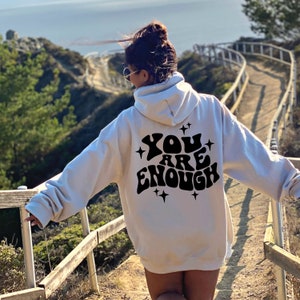 Puede incluir: Sudadera con capucha blanca con texto negro en la espalda que dice "YOU ARE ENOUGH" con estrellas alrededor del texto.