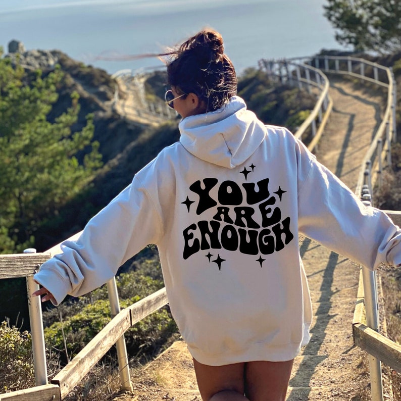 Puede incluir: Sudadera con capucha blanca con texto negro en la espalda que dice "YOU ARE ENOUGH" con estrellas alrededor del texto.