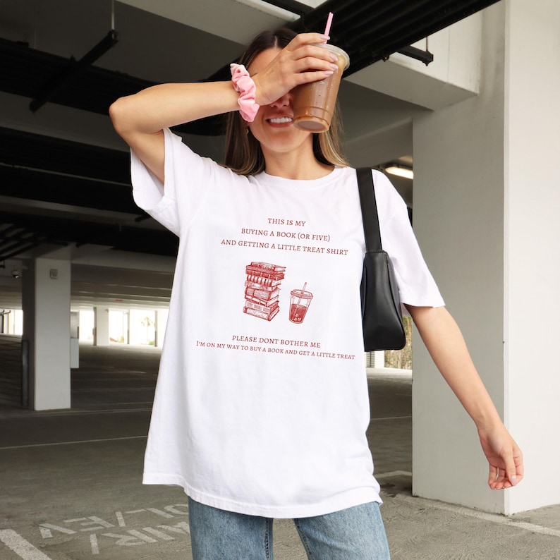 May include: White t-shirt with the text "THIS IS MY BUYING A BOOK (OR FIVE) AND GETTING A LITTLE TREAT SHIRT" in red. Below the text is a graphic of a stack of books and a drink. The shirt also says "PLEASE DONT BOTHER ME I'M ON MY WAY TO BUY A BOOK AND GET A LITTLE TREAT."