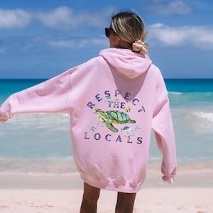 Könnte beinhalten: Rosa Kapuzenpullover mit einem Meeresschildkröten-Motiv und dem Text "Respect the Locals" auf der Rückseite.
