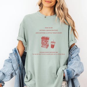 May include: A light green t-shirt with red text and graphics. The text reads "THIS IS MY BUYING A BOOK (OR FIVE) AND GETTING A LITTLE TREAT SHIRT. PLEASE DONT BOTHER ME I'M ON MY WAY TO BUY A BOOK AND GET A LITTLE TREAT." The graphic shows a stack of books and a drink.