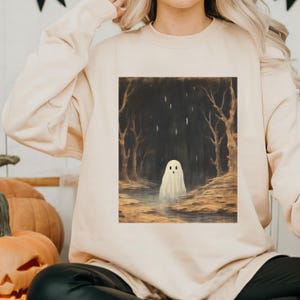Puede incluir: Una sudadera de color crema con un estampado gr&aacute;fico de un fantasma blanco en un bosque oscuro. El fantasma tiene una leve sonrisa y est&aacute; de pie en un charco de agua.