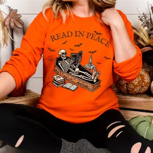 May include: Orange long-sleeved sweatshirt with a skeleton reading a book in a coffin. The text "Read in Peace" is printed on the shirt.