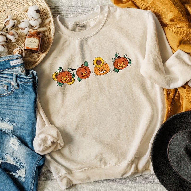 May include: A cream coloured sweatshirt with a graphic of four orange pumpkins with jack-o-lantern faces and green stems. The pumpkins are arranged in a row and have sunflowers and leaves around them.