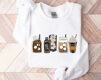 May include: White sweatshirt featuring a graphic of five different iced coffee drinks. The drinks include a mason jar, a glass bottle, a glass jar, a mason jar, and a takeaway cup. The text on the bottle reads "Cold Brew".