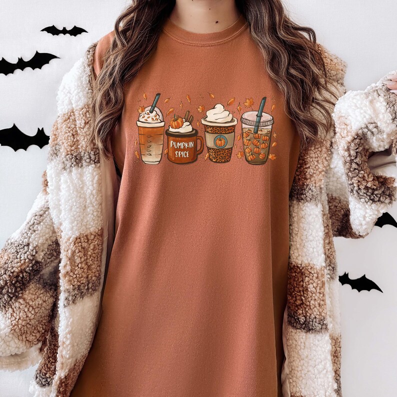 May include: A brown long-sleeved shirt with a graphic of four fall-themed coffee drinks. The drinks are labeled "Pumpkin Spice" and have fall-colored leaves and pumpkins around them.