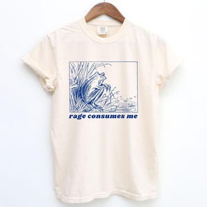 May include: A cream-colored t-shirt with a blue illustration of a frog in a pond. The text "rage consumes me" is printed in blue below the image.