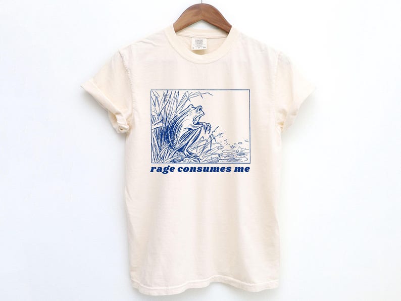 May include: A cream-colored t-shirt with a blue illustration of a frog in a pond. The text "rage consumes me" is printed in blue below the image.