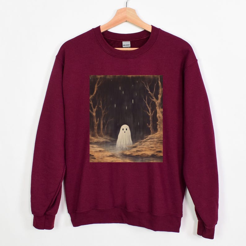 Puede incluir: Una sudadera color burdeos con un estampado gr&aacute;fico de un fantasma blanco en un bosque oscuro. El fantasma tiene una cara peque&ntilde;a y redonda y est&aacute; de pie en un charco de agua. El bosque es oscuro y misterioso, con &aacute;rboles altos y algunas hojas que caen.