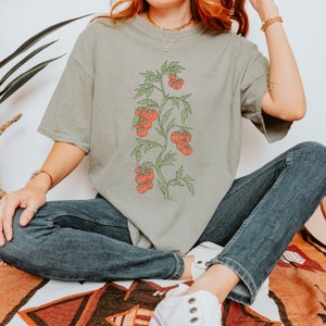May include: A woman wearing a light green t-shirt with a graphic of a tomato plant with red tomatoes. She is wearing blue jeans and white sneakers.