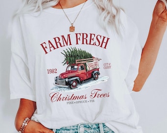 May include: White t-shirt with a red truck carrying a Christmas tree. The text on the shirt reads "Farm Fresh 1982 Christmas Trees Pine Spruce Fir"