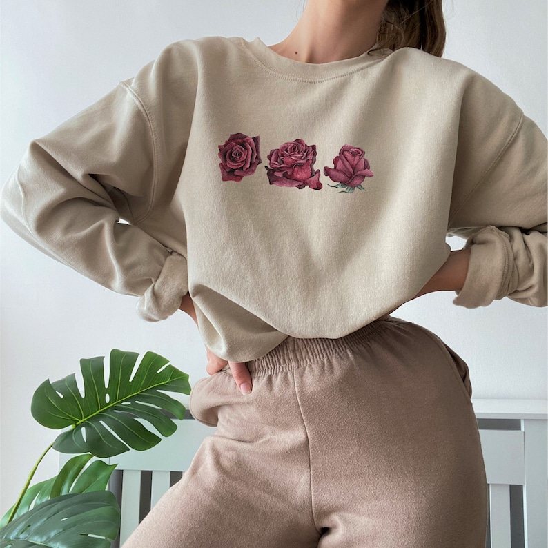 K&ouml;nnte beinhalten: Ein beiger Pullover mit Rundhalsausschnitt und langen &Auml;rmeln zeigt einen grafischen Druck mit drei roten Rosen. Der Pullover wird mit einer braunen Hose kombiniert.