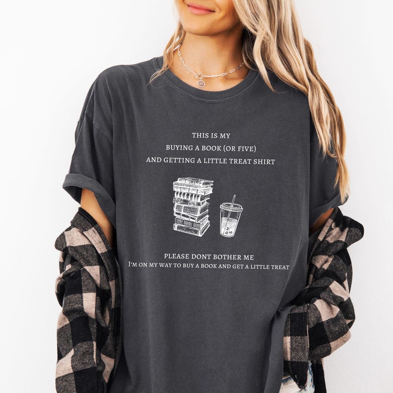 May include: Dark gray t-shirt with white text and illustrations. The shirt reads "THIS IS MY BUYING A BOOK (OR FIVE) AND GETTING A LITTLE TREAT SHIRT" with a stack of books and a drink graphic. The bottom text says "PLEASE DONT BOTHER ME I'M ON MY WAY TO BUY A BOOK AND GET A LITTLE TREAT."