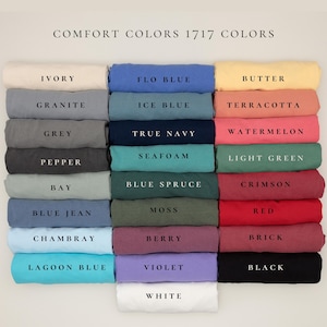 May include: A display of folded Comfort Colors 1717 t-shirts in various colors, including Ivory, Flo Blue, Butter, Granite, Ice Blue, Terracotta, Grey, True Navy, Watermelon, Pepper, Seafoam, Light Green, Bay, Blue Spruce, Crimson, Blue Jean, Moss, Red, Chambray, Berry, Brick, Lagoon Blue, Violet, Black, and White.