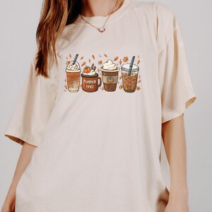 May include: A beige t-shirt with a graphic of four different fall-themed coffee drinks. The drinks are decorated with fall leaves and pumpkins. The text "Pumpkin Spice" is printed on the shirt.